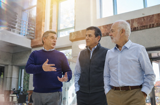 Three professionally dressed men walking together inside a modern office building with large windows and natural light. One man gestures while speaking, and the other two listen attentively as they engage in conversation.