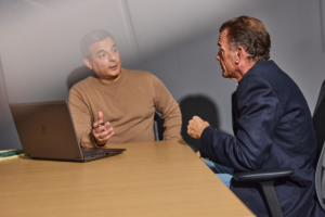 Two business partners sit across from each other at a wooden conference table engaged in an intense discussion. One man, seated beside an open laptop, gestures with his hand while explaining a point, while the other leans forward attentively in a chair, appearing to respond or challenge the idea. The setting appears to be a professional office environment, suggesting a serious conversation about a business decision, negotiation, or partnership matter.