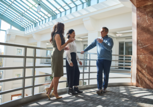 Professional setting: Three business professionals stand in a bright, modern office corridor with a glass ceiling, engaged in an animated discussion. One man gestures while speaking, as two women listen and respond, suggesting a collaborative conversation or strategic meeting.