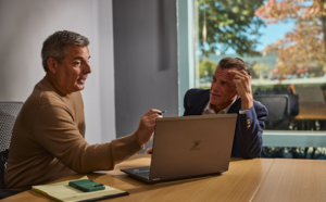 Two business professionals seated at a conference table engaged in a serious discussion, one pointing at a laptop while the other listens with a concerned expression, suggesting a high-stakes or problem-solving conversation.