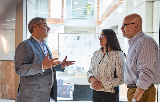 Three professionals engaged in a focused discussion inside a modern office space, with one man gesturing as he speaks while a woman and another man listen attentively, suggesting a serious business or advisory conversation.