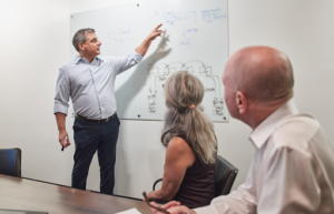 A professional business meeting scene where Len stands at a whiteboard, pointing to a diagram as he explains a concept. Two colleagues—a woman and a man—sit at a conference table, attentively listening and looking toward the board. The setting is a clean, modern office, suggesting a strategic discussion or planning session.