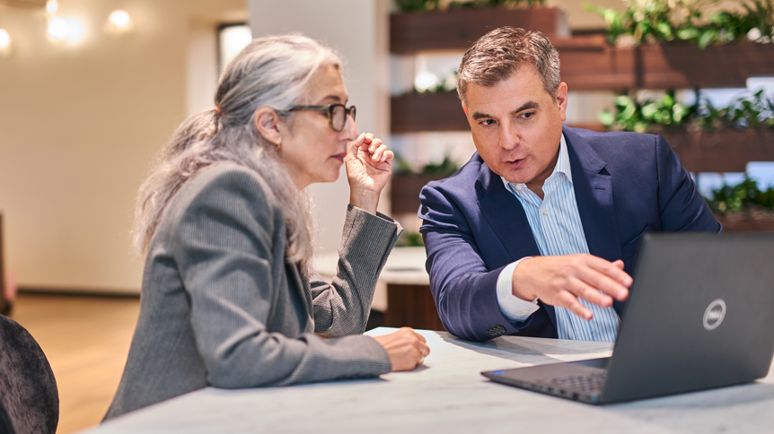 Professional setting with two individuals engaged in a focused discussion—one person gestures toward a laptop screen while the other listens attentively—suggesting a collaborative conversation around financial planning, strategy, or business decision-making.