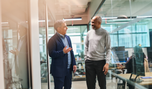 Two business professionals walking through a modern glass-walled office, engaged in conversation—one in a navy suit gesturing as he speaks, the other in a grey sweater listening and smiling, with desks and coworkers visible in the background.