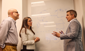 Three professionals engaged in a discussion during a meeting beside a glass whiteboard covered with diagrams and handwritten notes. A man in a gray suit gestures while speaking to a woman and another man who listen attentively in an office setting.