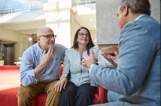 A professional meeting between three adults in a modern office lounge setting. A man and woman are seated together on a red couch, attentively listening to another man in a blue blazer who is speaking and gesturing with his hands. The atmosphere appears collaborative and conversational, suggesting a consultation, advisory meeting, or business discussion in a bright contemporary workspace.