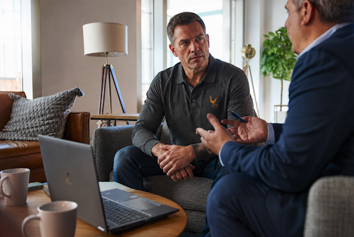 Two business professionals seated in a modern office lounge engaged in a serious discussion. One man in a dark polo shirt listens attentively while the other, dressed in a blue suit, gestures as he speaks. A laptop sits open on a coffee table between them, with coffee mugs and office décor visible in the background, creating a professional and collaborative atmosphere.