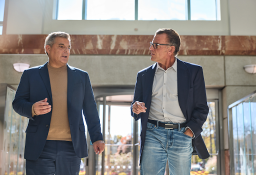 Two professionally dressed men walking side by side outside a modern office building, engaged in conversation. One wears a navy blazer with a tan sweater, while the other wears a navy blazer with a white shirt and jeans. Natural light streams through large glass windows behind them, suggesting a business discussion in a corporate setting.