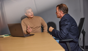 Two business professionals sit across from each other at a conference table during a meeting. One man gestures while speaking beside an open laptop, while the other listens attentively from an office chair. The setting is a modern office with neutral gray walls, conveying a focused discussion about business, legal, or financial matters.