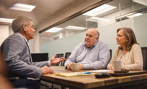 An older couple sits across from a professional advisor in a modern conference room, discussing financial or estate planning matters. The advisor gestures while speaking, and the couple listens attentively with serious expressions. Coffee mugs, documents, and office furnishings create a professional setting, suggesting a consultation about wealth preservation, inheritance planning, or family legacy issues.