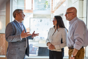 Three professionals engaged in conversation inside a modern office building with large windows and natural light. A man in a gray blazer gestures while speaking to a woman in a white patterned jacket and a man in a striped shirt. The group appears to be discussing business or legal matters in a collaborative and professional setting.
