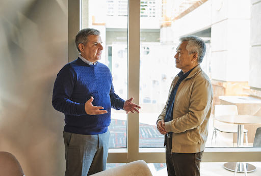 Two professionally dressed men stand in conversation inside a modern office space near large floor-to-ceiling windows. One man, wearing a blue sweater, gestures with his hands while speaking, while the other, dressed in a tan jacket, listens attentively. Natural light fills the room, creating a bright and professional atmosphere, with contemporary furnishings visible in the background.