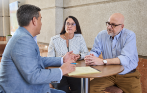 An attorney in a blue suit meets with a married couple seated at a small table in a professional office setting. The attorney gestures while explaining estate planning options as the couple listens attentively, discussing trusts, wealth preservation, and long-term legacy planning.