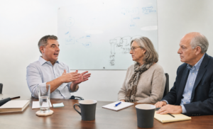 A man in a light blue dress shirt gestures while speaking to two older adults seated across from him at a conference table during a professional estate planning consultation. The woman holds a notebook and pen, while the man has a yellow legal pad in front of him. Coffee mugs, a glass of water, and a whiteboard with handwritten notes in the background create the setting of a thoughtful legal or financial planning discussion in a modern office.