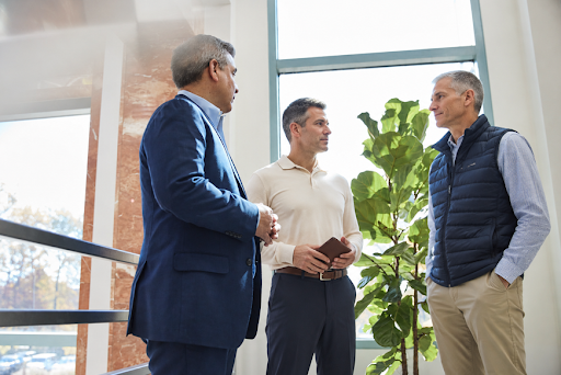 Three business partners or advisors stand together in a bright, modern office discussing business matters. One man holds a notebook while the others listen attentively, illustrating a professional conversation about business succession, partnership planning, or buy-sell agreement valuation. Large windows and indoor greenery create a welcoming corporate environment.