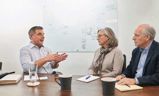 A man in a light blue dress shirt gestures while speaking to two older adults seated across from him at a conference table during a professional estate planning consultation. The woman holds a notebook and pen, while the man has a yellow legal pad in front of him. Coffee mugs, a glass of water, and a whiteboard with handwritten notes in the background create the setting of a thoughtful legal or financial planning discussion in a modern office.