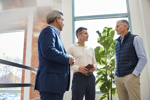 Three professional men stand in a bright, modern office having a conversation, representing business owners and advisors discussing estate planning and strategic planning before the sale of a business.