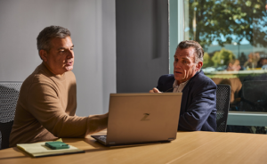 Two business professionals sit across from each other at a conference table, reviewing information on a laptop during a meeting. One gestures toward the screen while the other listens attentively, suggesting a discussion about business strategy or planning in a modern office with large windows and natural light.