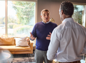 Two men having a professional conversation in a bright office, with one man gesturing while speaking near large windows and a leather sofa.