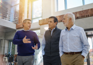 Three mature businessmen having a conversation in a bright, modern office building, with one man gesturing as the others listen.