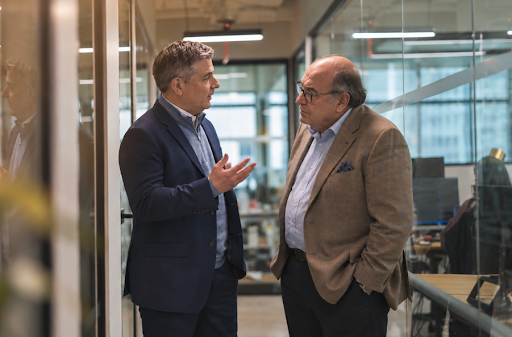 Two men in business attire having a professional conversation in a modern office hallway, with one man gesturing as the other listens.