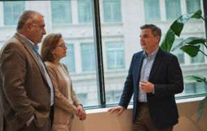 Three professionals having a conversation beside large office windows, with one man speaking while the other man and woman listen.